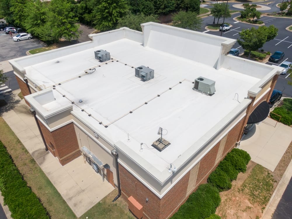 Aerial view of a single-story brick and stucco commercial building with a white flat roof, multiple HVAC units, and parking areas with some trees and cars visible in the background.