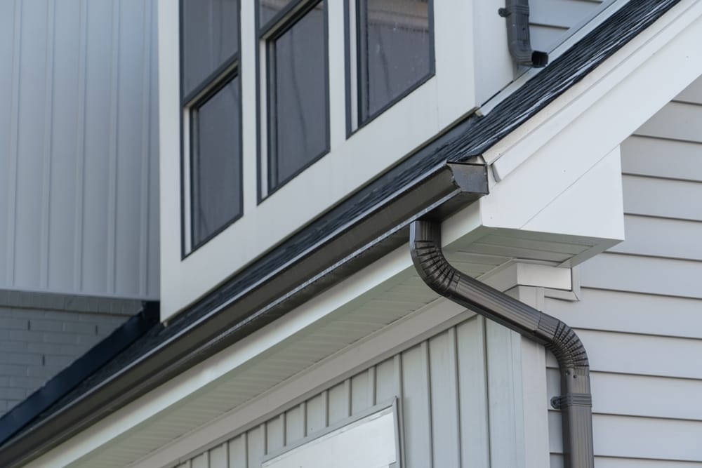 Close-up view of a house exterior showing a section of the roof, a downspout, and siding with a window above.