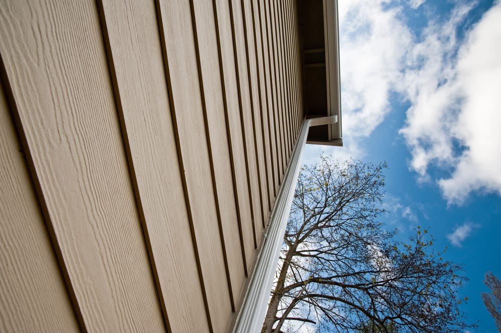 Upward view of the side of a building with beige siding, adjacent to a leafless tree, under a partly cloudy blue sky.